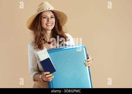 Strandurlaub. Lächelnde elegante 40-jährige Frau in weißer Bluse und Shorts vor beigefarbenem Hintergrund mit Sommerhut und Trolley-Tasche. Stockfoto
