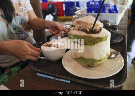 Frau genießt erfrischendes tropisches Kokosnuss-Dessert und heißen Latte im Café. Dieses einzigartige Getränk in Kokosnussschale und köstliches Getränk auf Tablett bieten Rela Stockfoto
