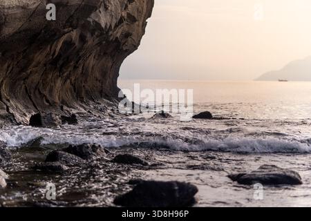 Flacher Blick auf Meereswellen, die in der Nähe einer dramatischen, geschwungenen Klippe auf dunkle Felsen treffen. Ruhige Meereslandschaft mit sanftem Horizontlicht in der Dämmerung. Stockfoto