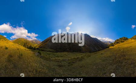 Ein weites Panorama fängt ein Bergtal mit sanften Hängen ein, die mit gelb-grünem Gras bedeckt sind. Die Sonne scheint hell hinter einem zentralen Berggipfel und erzeugt Strahlen am klaren blauen Himmel mit verstreuten weißen Wolken. Stockfoto