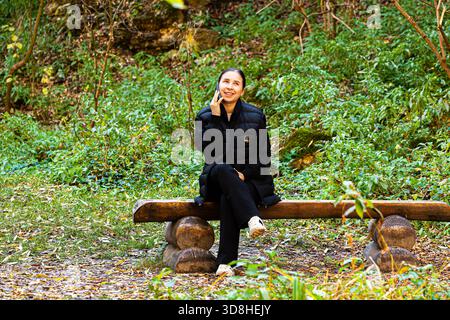Frau Freiberuflerin trägt Leinenkleid, sitzt auf einem Stuhl im Gewächshaus, ruht aus, benutzt Smartphone und trinkt Tee aus einer Tasse im Einklang mit den Pflanzen Stockfoto