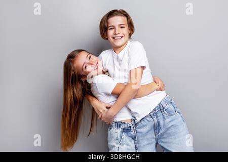 Fröhliche Kinder, die sich im Studio umarmen und lächeln, verspielte Freunde, die Spaß miteinander genießen, in lässigen weißen T-Shirts vor sauberem grauem Hintergrund Stockfoto