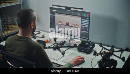 Techniker mit tätowierten Tests von FPV Drohnenelektronik, Anschließen von Drähten und Prüfen der Flugparameter der Drohne auf dem Computermonitor. Konzept Technische Kalibrierung und Qualitätssicherung von FPV Quadcoptern. Stockfoto
