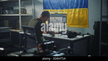 Ukrainischer Ingenieur mit Tattoos überprüft moderne FPV Drohne mit Computer, mit ukrainischer Flagge hängt an der Wand hinter dem Menschen. Konzept der nationalen Bemühungen im Bereich der Verteidigungstechnologien und der Fertigung. Stockfoto