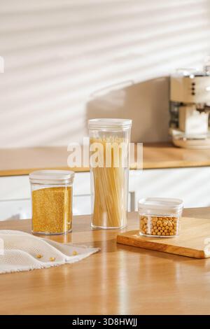 Minimalistische Küchenkomposition mit drei durchsichtigen Lebensmittelbehältern gefüllt mit Spaghettihirse und Sojabohnen auf Holztisch. Stockfoto