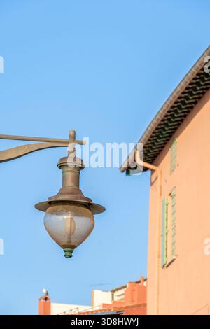 Nahaufnahme einer staubigen Vintage-Straßenlaterne vor einem hellblauen Himmel, mit der Ecke eines lachsrosa Gebäudes und grünen Fensterläden sichtbar Stockfoto