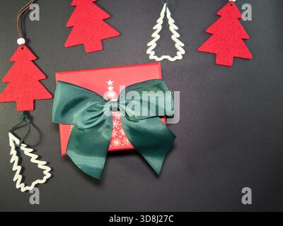 Festliche Geschenkbox und Weihnachtsdekoration – Rote Geschenkbox mit grüner Schleife und Band, umgeben von festlichen Weihnachtsbaumschmuck auf dunklem Hintergrund. Leistung Stockfoto