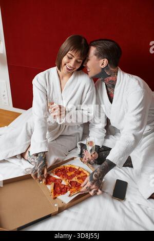 Ein junges Paar teilt einen freudigen Moment, lacht und genießt Pizza, während es in Hotelmantel gekleidet ist. Stockfoto