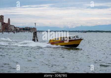 Gelbes und rotes Rettungsboot, das über die venezianische Lagune bei Murano mit historischen Gebäuden und fernen Bergen im Hintergrund rastet, Venedig, Stockfoto