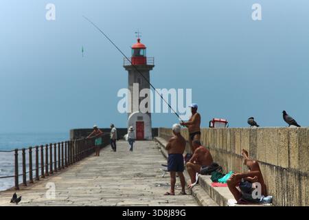 Die Fischer entspannen sich und fischen auf einem sonnigen Pier mit einem hoch im Hintergrund stehenden Leuchtturm, der eine ruhige Küstenszene zeigt Stockfoto