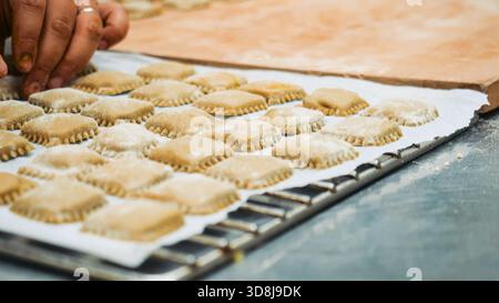 Arbeiterhände arrangieren Reihen hausgemachter, ungekochter Ravioli, die mit Mehl bestäubt sind, auf einem mit Pergament ausgelegten Metalltablett in einer professionellen Küche, wobei Handwerker gezeigt werden Stockfoto