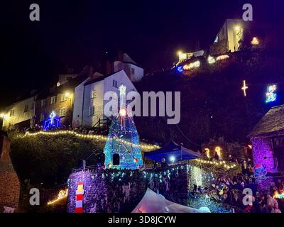 Menschenmassen versammeln sich um den Weihnachtsbaum im Hafen, während das RNLI-Weihnachtslicht in Clovelly mit beleuchteten Hütten und Dekorationen eingeschaltet wird. Stockfoto