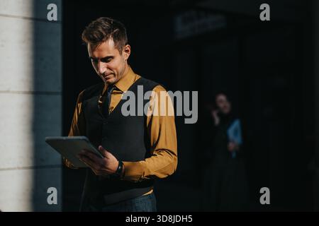 Geschäftsmann steht draußen, konzentriert auf ein Tablet, trägt Senfhemd und Weste für eine professionelle Lifestyle-Stimmung Stockfoto