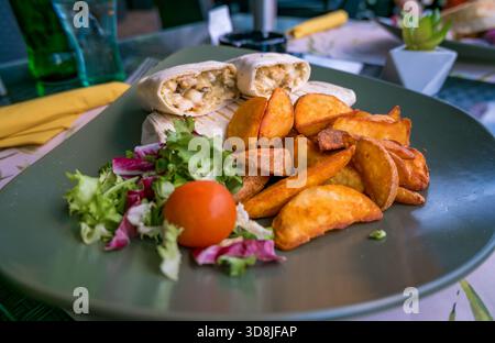 Ein Teller mit zwei Wraps, goldenen Kartoffelscheiben und einem frischen Salat mit einer Kirschtomate. Stockfoto