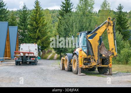 Ein gelber Baggerlader, der auf einer Schotterstraße in der Nähe eines weißen Lkws geparkt ist. Stockfoto