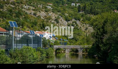 Solarpaneele in Gebäuden in der Nähe eines Flusses, umgeben von grünen Hügeln und Bäumen. Stockfoto