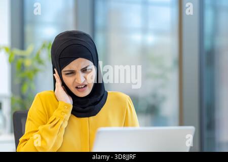 Muslimische Frau, die einen Hijab trägt und sich auf ihren Laptop konzentriert, während sie schmerzhafte Ohrenschmerzen oder Kopfschmerzen erlebt, während sie ihr Ohr mit einem schmerzhaften Ausdruck in einer Büroumgebung berührt Stockfoto
