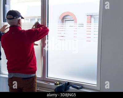 Älterer Arbeiter, der durchscheinende Privatsphärenfolie auf ein Fenster aufträgt Stockfoto