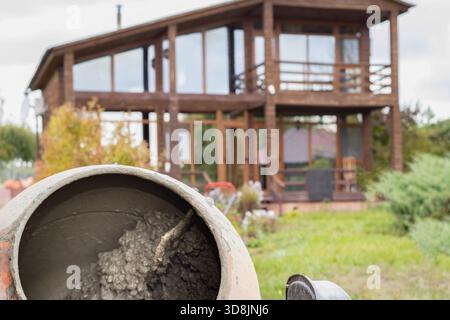 Alter Zementmischer mit getrocknetem Beton, der während der Bauarbeiten in der Nähe eines Holzhauses steht Stockfoto