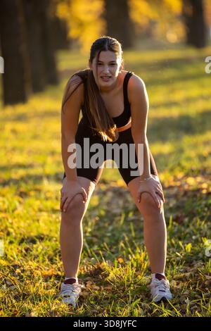 Eine junge Frau macht im Herbst eine Pause vom Joggen in einem Park. Sie beugt sich nach vorne und atmet im warmen Sonnenlicht. Stockfoto