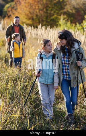 Eine vierköpfige Familie spaziert durch die sonnige Wiese, genießt ihre Wanderung, während sie Wanderstöcke trägt und tagsüber zusammen lächelt Stockfoto