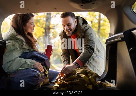 Ein Paar genießt es, an einem sonnigen Herbsttag, umgeben von bunten Blättern und Natur, ihre Ausrüstung im hinteren Teil ihres Autos vorzubereiten. Stockfoto