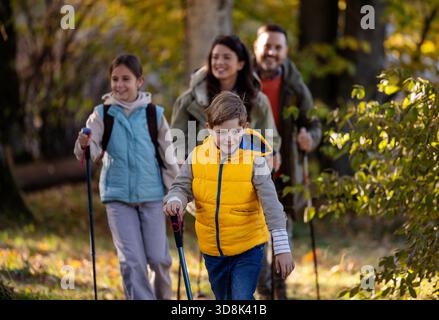Vierköpfige Familie spaziert durch den sonnigen Wald, genießt ihre Wanderung, während sie Wanderstöcke trägt und tagsüber zusammen lächelt Stockfoto