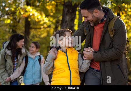 Vierköpfige Familie spaziert durch den sonnigen Wald, genießt ihre Wanderung, während sie Wanderstöcke trägt und tagsüber zusammen lächelt Stockfoto