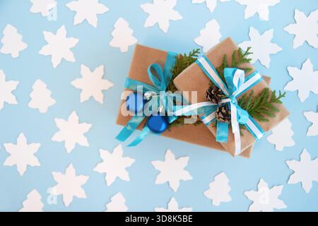 Zwei Geschenkboxen mit Bastelpapier, blauem und weißem Band und verzierten Tannenzweigen, blauen Weihnachtskugeln und Tannenzapfen auf blauem Hintergrund mit weißer Papiertanne und Stern, Ansicht von oben. Weihnachtsgeschenke. Stockfoto