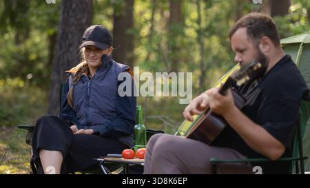 Bärtiger Mann spielt Gitarre für lächelnde Frau, während er im Wald campt, sitzt auf Klappstühlen neben Zelt und Lagerfeuer, mit Tomaten, Gurken und Stockfoto