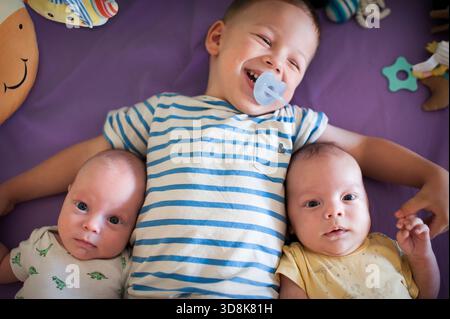 Ein glücklicher älterer Bruder mit einem Schnuller im Mund liegt mit seinen neugeborenen Zwillingsgeschwistern auf einer lila Decke. Bunte Spielzeuge sind überall verstreut Stockfoto