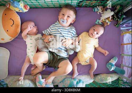 Ein glücklicher älterer Bruder mit einem Schnuller im Mund liegt mit seinen neugeborenen Zwillingsgeschwistern auf einer lila Decke. Bunte Spielzeuge sind überall verstreut Stockfoto