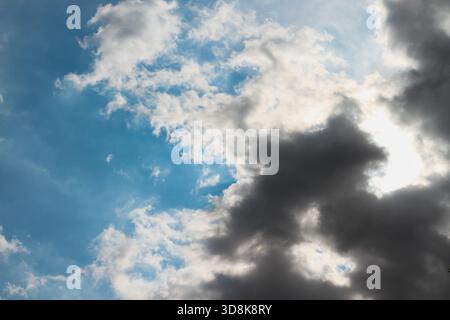 Die Sonne versteckt sich hinter einer Masse dunkelgrauer Wolken, mit Lichtstrahlen und Schatten auf blauem Himmel, als Hintergrund Stockfoto