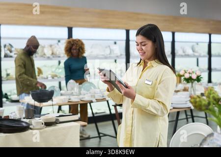 Junge Erwachsene Frau, die ein digitales Tablet benutzt, während sie in einem modernen Geschäft steht, lächelt und sich auf den Bildschirm konzentriert, Paar, das im Hintergrund Geschirr durchsucht, Einzelhandel Stockfoto