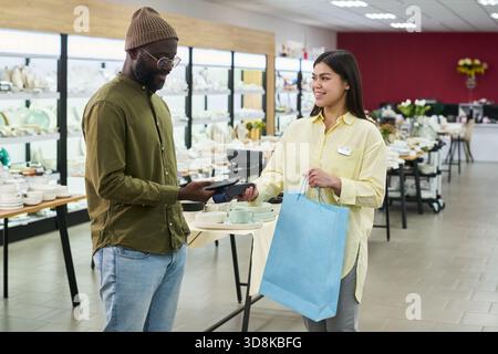 Kaukasische junge Erwachsene Frau hilft Schwarzer, junger, erwachsener Mann, der mit Kartenleser im Geschäft bezahlt, Mann mit elektronischem Gerät, während Frau mit Einkaufstasche und lächelndem Lächeln Stockfoto