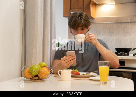Kaffee trinken und Telefon checken junger Mann, der am Küchentisch sitzt und warme Getränke genießt. Gesundes Frühstück. Wohnküche Lifestyle Stockfoto