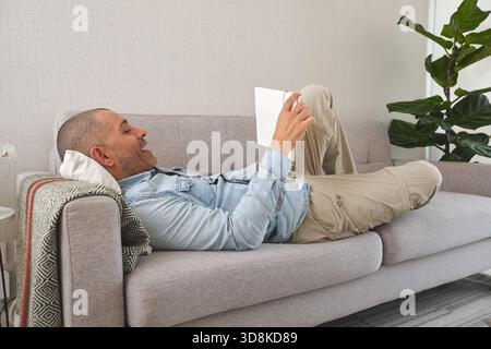 Ein glücklicher Mann, der gerne ein Buch liest, während er auf einem bequemen Sofa liegt Stockfoto