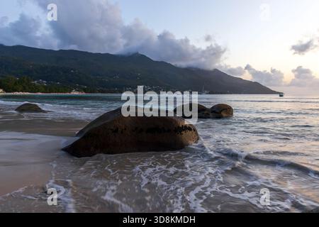 Seychellen Küstenszene mit glatten Felsen im Vordergrund, sanften Wellen, die über die Sandküste ziehen, üppig grünen Bergen im Hintergrund A Stockfoto