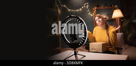 Weibliche Bloggerin packt die Box aus, nimmt Videos mit Smartphone auf, Ringlicht mit Stativ sagt, tut mir leid, weiß es nicht am späten Abend im Home Office. Onli-Konzept Stockfoto