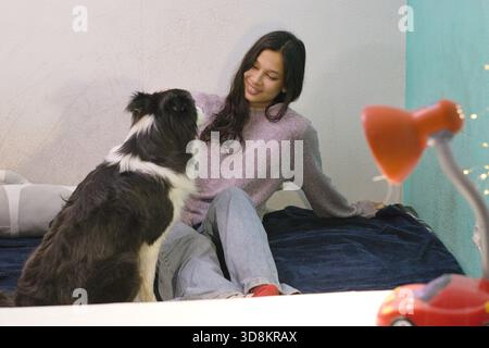 Junge Frau sitzt auf dem Bett, Border Collie schaut sie an, entspannte Indoor-Szene mit sanfter Beleuchtung, Interaktion mit Haustieren und Personen, perfekt für den Lifestyle, Haustier c Stockfoto