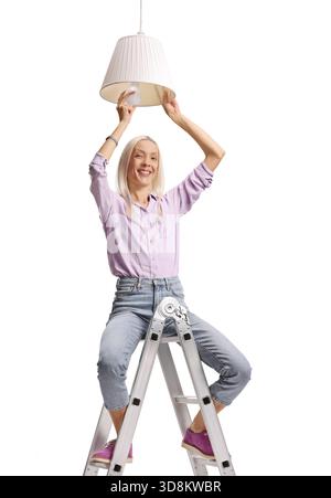 Frau auf einer Leiter, die eine Lampe auf eine Pendelleuchte auf weißem Hintergrund setzt Stockfoto