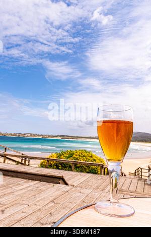 Erfrischendes Glas kaltes Bier auf einem Holztisch mit Blick auf einen wunderschönen Sandstrand, das blaue Meer und den bewölkten Himmel. Perfekter Sommerurlaub und Entspannung Stockfoto