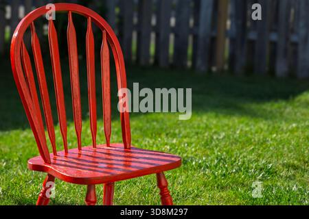 Ein leuchtend roter Holzstuhl auf einem sonnendurchfluteten grünen Rasen mit einem verschwommenen Zaun im Hintergrund. Stockfoto