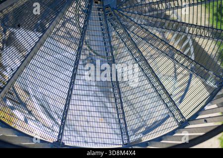 Abstrakte Ansicht einer blauen Metallspiraltreppe mit Sonnenlicht, die geometrische Schatten und Reflexionen auf den Boden darunter wirft. Stockfoto