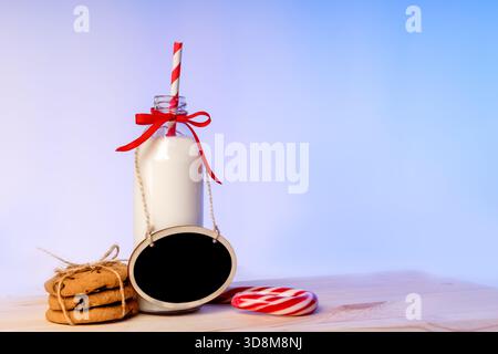 Eine Flasche Milch mit rotem Band und Strohhalm, ein Stapel Kekse und ein leeres schwarzes Miniatur-Schild, das an Garn hängt, neben einem rot-und-wh Stockfoto