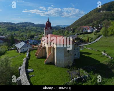 Blick aus der Vogelperspektive auf die markante Kirche mit rotem Dach, umgeben von sanften grünen Hügeln und malerischen Dorfhäusern, eine ruhige Mischung aus Natur und Architektur, Lubietov Stockfoto