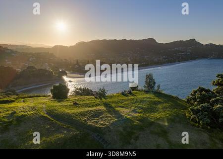 Aus der Vogelperspektive auf die Sonne, die die fernen Hügel küsst, und den ruhigen Strand, der auf das türkisfarbene Meer trifft, Hahei, Waikato Region, Neuseeland. Stockfoto