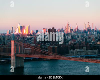Blick aus der Vogelperspektive auf die Brooklyn Bridge, die sich über den East River in Richtung der beleuchteten Skyline von Manhattan erstreckt, getaucht in das warme Licht des Sonnenuntergangs, N Stockfoto