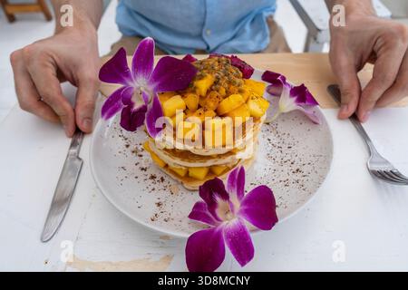 Leckere Pfannkuchen mit tropischen Früchten auf einem Teller auf einem Tisch in einem Café, Nahaufnahme. Gesundes Frühstück. Leckere Pfannkuchen mit Mango, Drachenfrucht und Pass Stockfoto