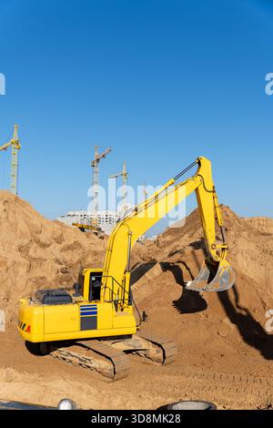 Ein großer gelber Bagger arbeitet auf einer Baustelle, umgeben von Schmutzhügeln, mit Turmkränen und unfertigen Gebäuden im Hintergrund unter einem Stockfoto